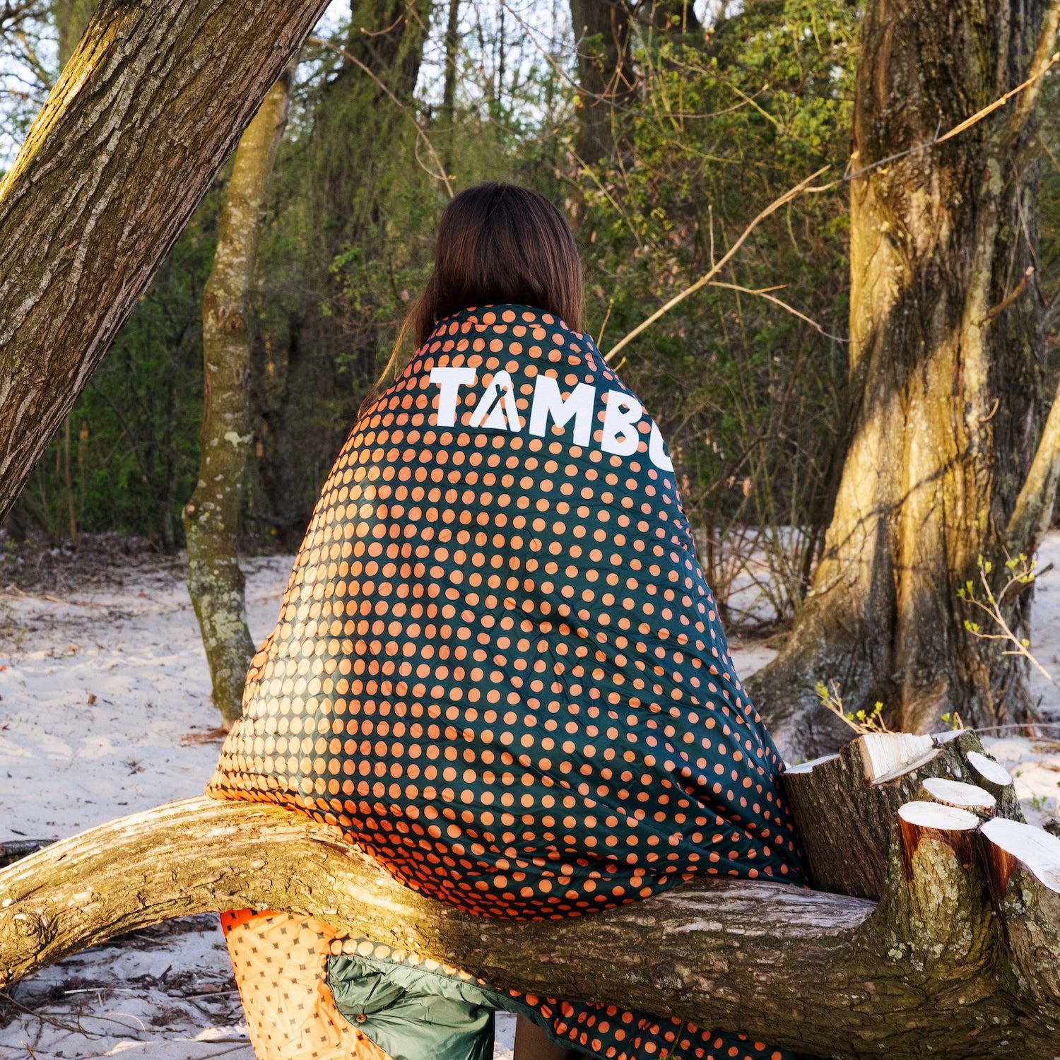 Person sitzt auf einem Baumstamm im Wald, umhüllt von einer dunkelgrünen Campingdecke mit orangefarbenen Punkten und weißem 'TAMBU' Schriftzug