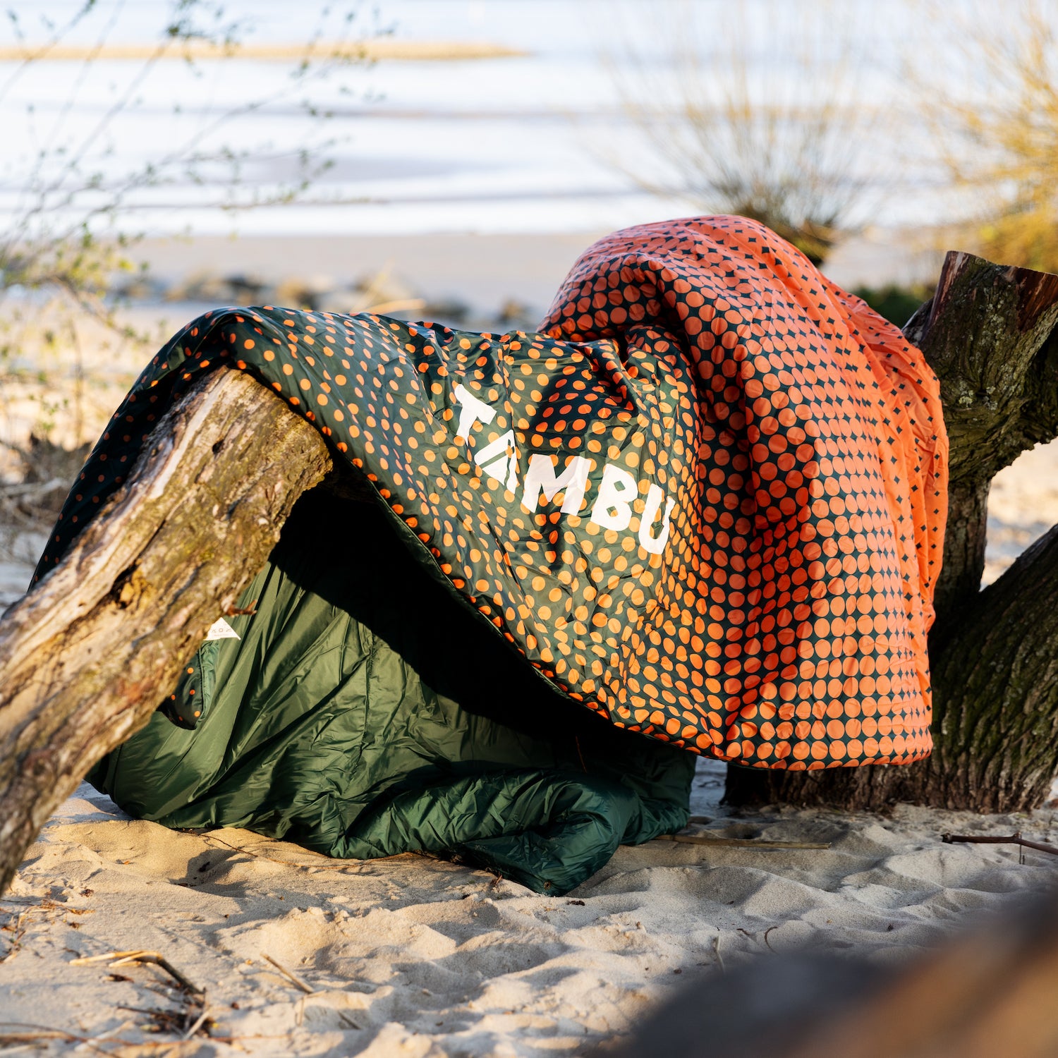 Ultraleichte Campingdecke KAMBALA von Tambu Outdoor mit orange-schwarzem Punktmuster auf Sand zwischen Baumstämmen drapiert