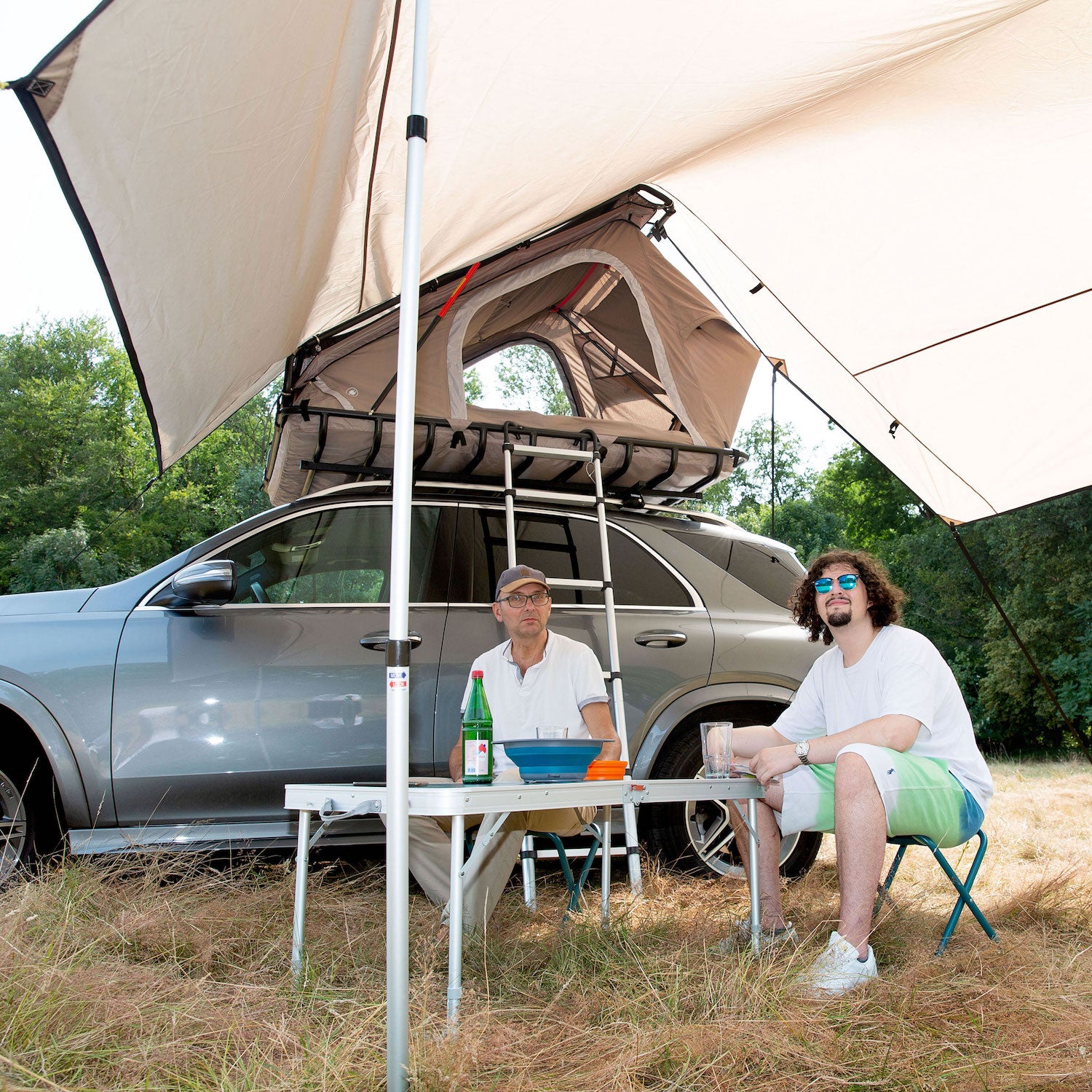 Zwei Männer sitzen unter dem YANO Sonnensegel von Tambu Outdoor neben einem grauen Auto mit geöffnetem Dachzelt auf Campingstühlen an einem Klapptisch mit Getränken und Geschirr im Freien