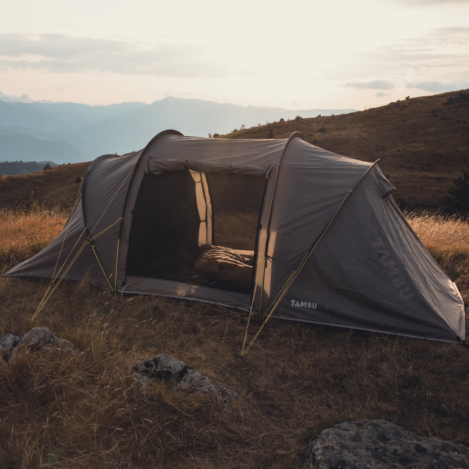 AGNIKUNDA 4-Personen Trekkingzelt von Tambu Outdoor, braun, Eingänge geöffnet, auf einer Wiese mit Bergen im Hintergrund bei Sonnenuntergang