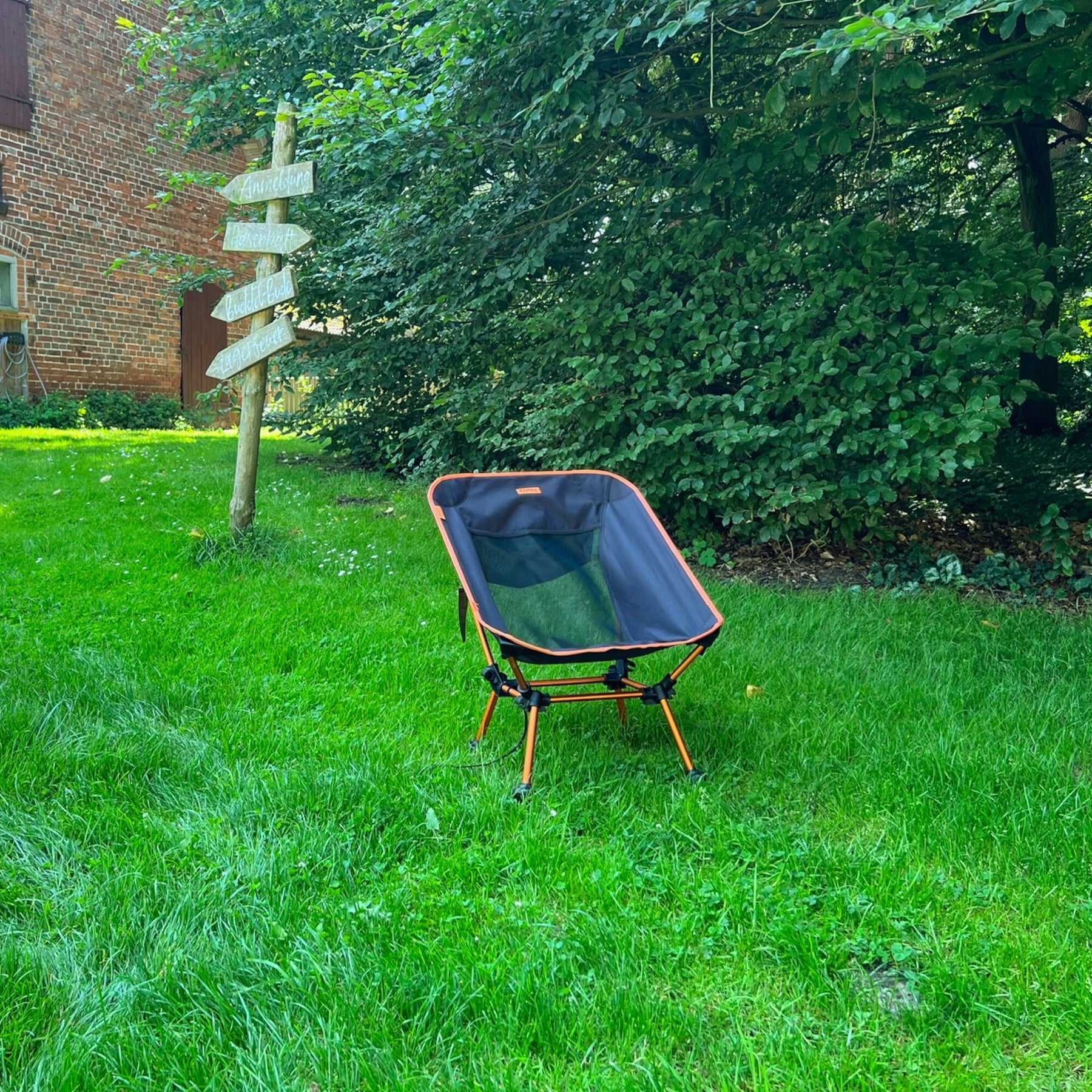 Leichter Campingstuhl BATASA von Tambu Outdoor in Schwarz mit orangefarbenem Rahmen, steht auf grünem Gras vor Büschen und einem Holzwegweiser