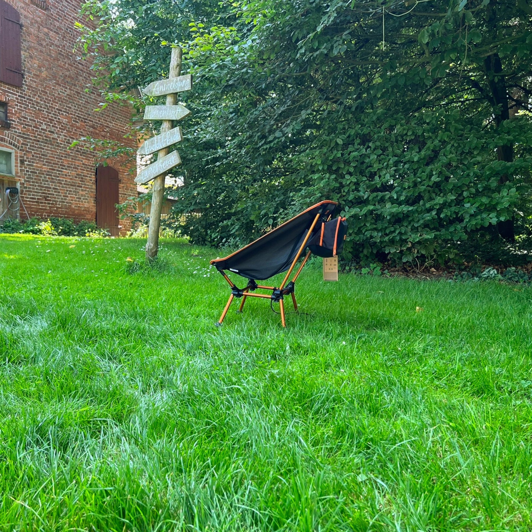 Leichter Campingstuhl BATASA von Tambu Outdoor in schwarz mit orange, mit zwei Sitzhöhen, steht auf grünem Gras vor einem Holzwegweiser und Bäumen