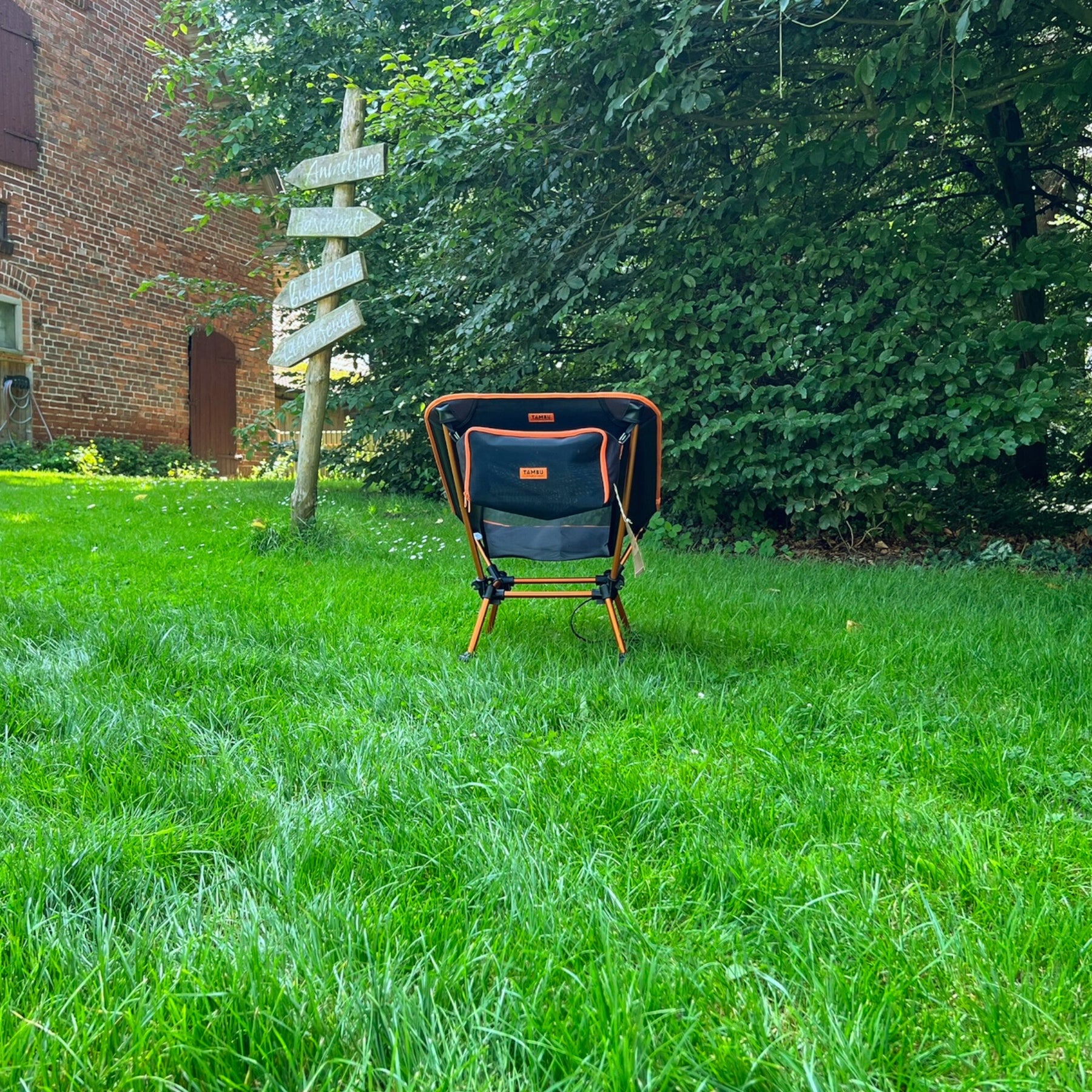 Schwarzer und oranger leichter Campingstuhl BATASA von Tambu Outdoor mit zwei Sitzhöhen steht auf grünem Gras vor einem Wegweiser und Bäumen