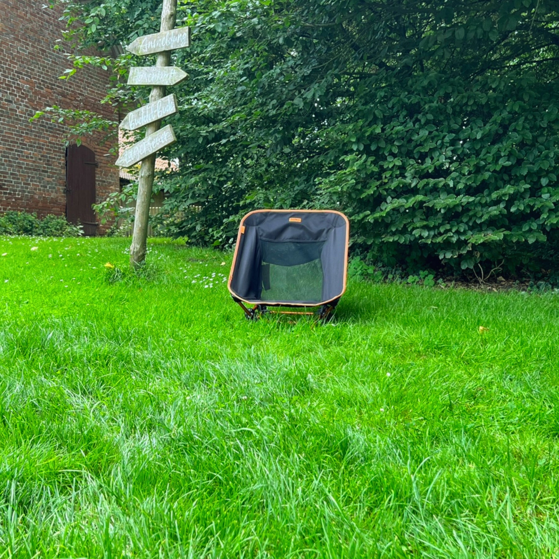 Schwarzer und oranger leichter Campingstuhl BATASA von Tambu Outdoor mit zwei Sitzhöhen auf grünem Gras vor Baum und Holzwegweiser