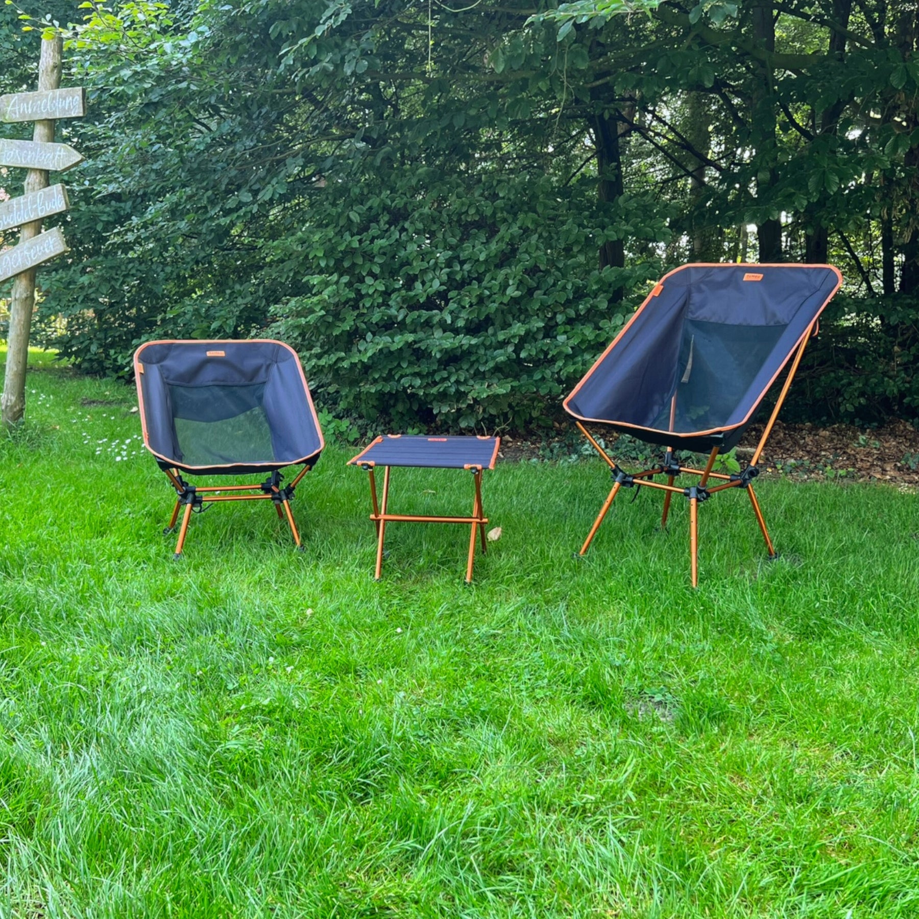 Zwei schwarze und orange Campingstühle von Tambu Outdoor mit unterschiedlichen Sitzhöhen und ein kleiner passender Tisch auf grünem Gras vor Bäumen