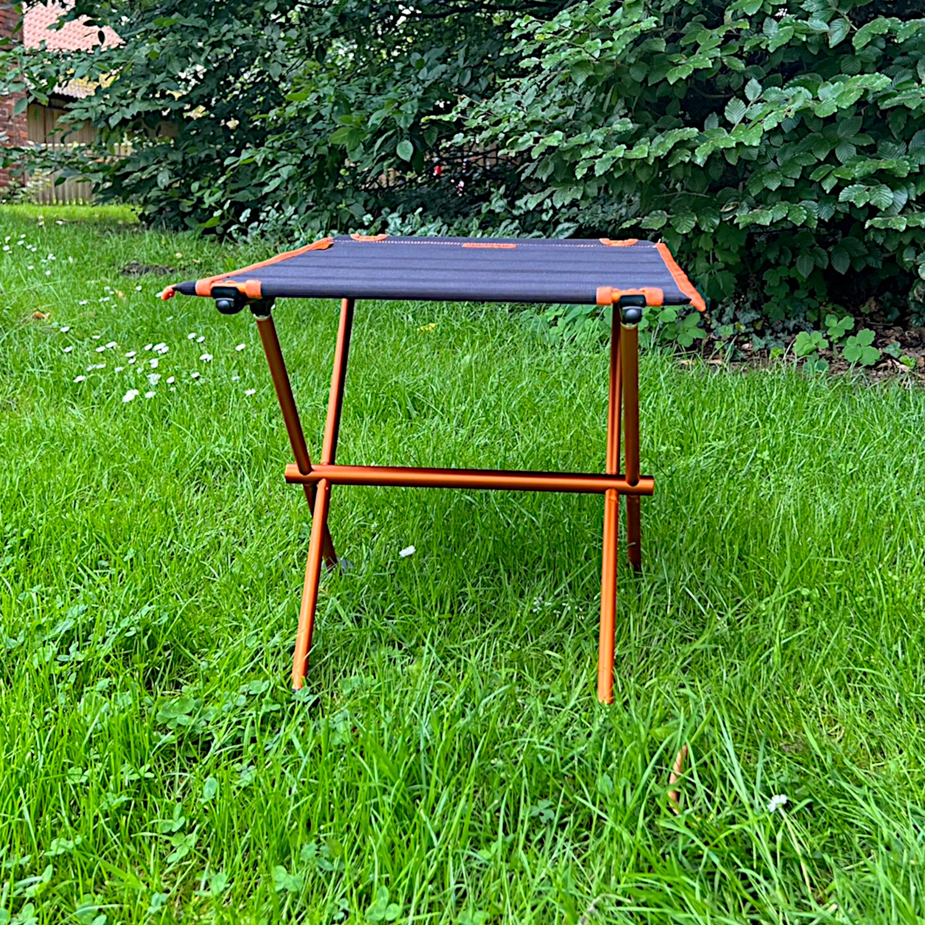 Leichter Campingtisch BATISTA von Tambu Outdoor mit schwarzer Tischplatte und orangefarbenem Aluminiumgestell auf grünem Gras