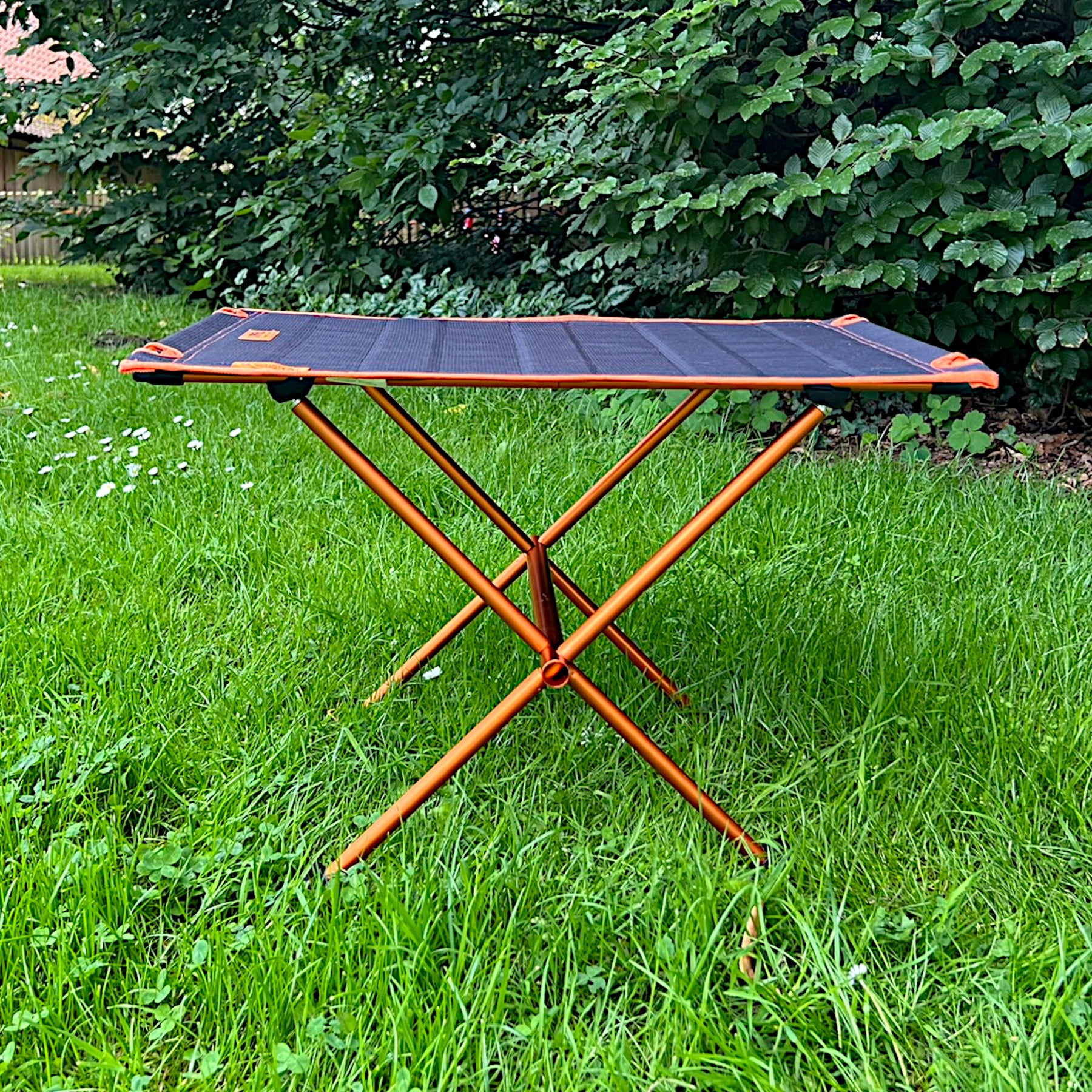 Leichter Campingtisch BATISTA von Tambu Outdoor mit schwarzer Tischplatte und orangefarbenem Gestell auf grünem Gras