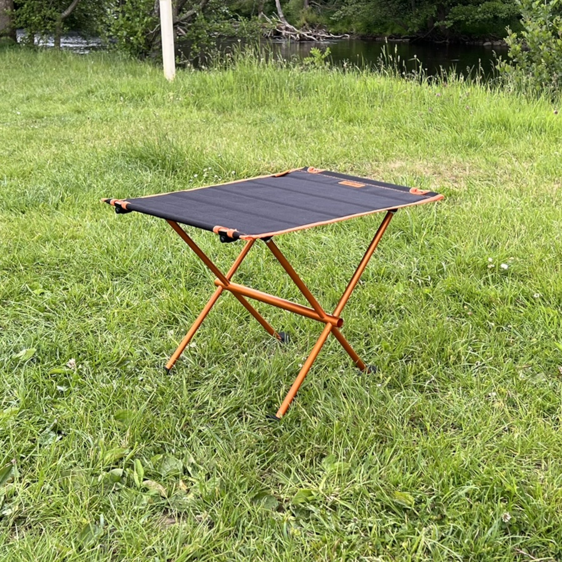 Leichter Campingtisch BATISTA von Tambu Outdoor mit schwarzer Aluminiumplatte und orangefarbenem Gestell auf Wiese