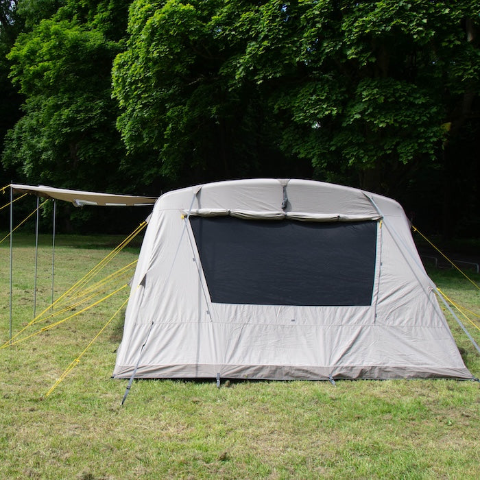 BISALA 5 Familien Tunnelzelt AIR von Tambu Outdoor in beige mit großem Fenster und Sonnendach auf grüner Wiese vor Bäumen