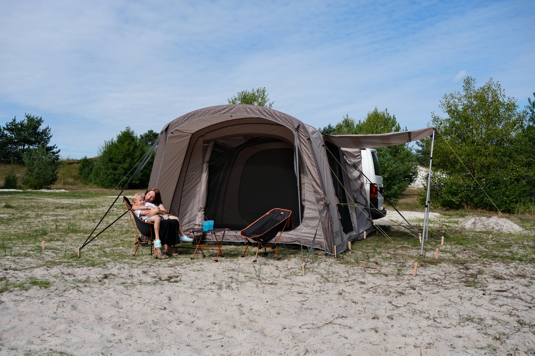 Beiges BISALA VAN Zelt von TAMBU Outdoor an einem weißen Van, mit zwei Personen auf Campingstühlen davor, auf sandigem Boden mit Büschen im Hintergrund.