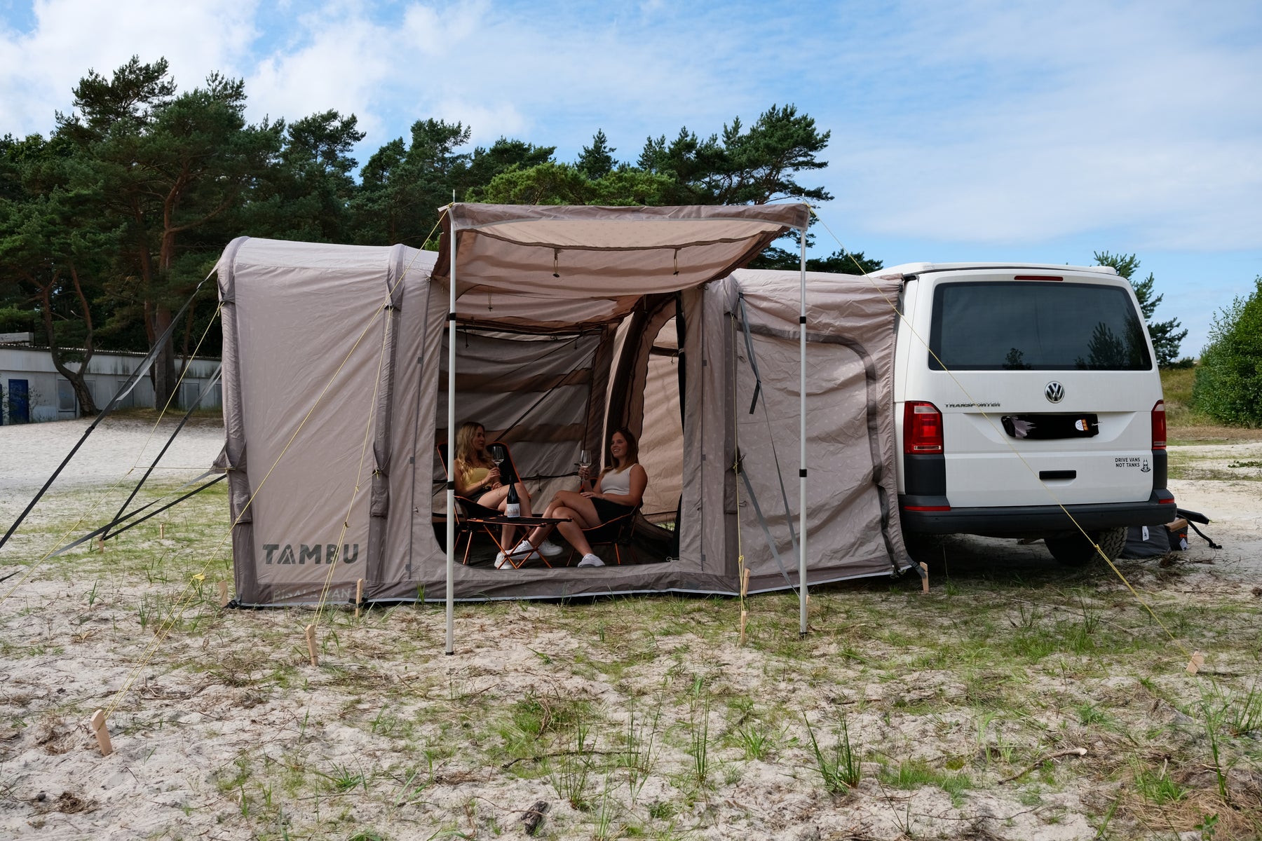 Beiges BISALA VAN von TAMBU Outdoor als Anbau am weißen VW-Bus, zwei Personen sitzen unter dem Vorzelt auf Campingstühlen.