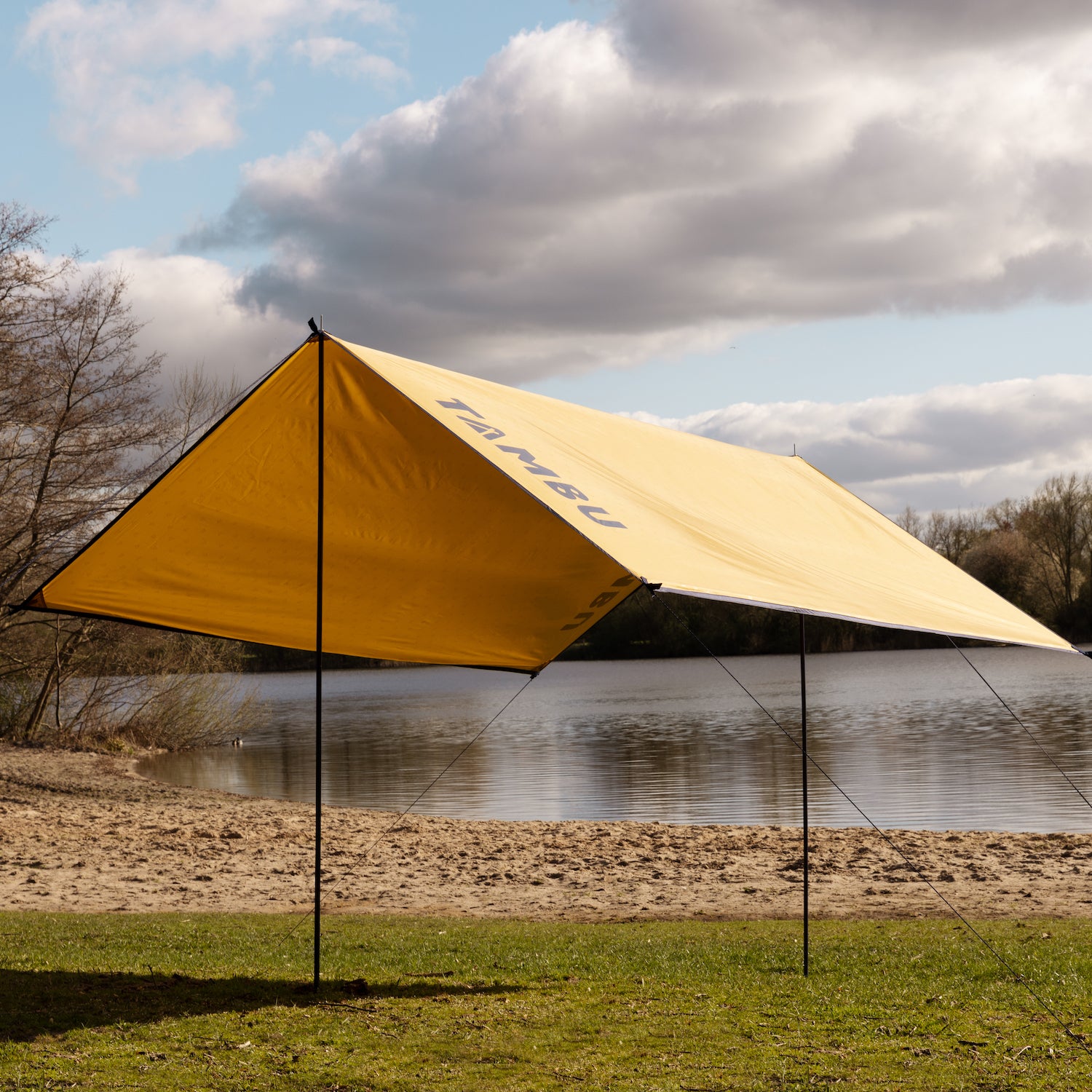 Gelbes multifunktionales Sonnensegel CHADA von Tambu Outdoor am Seeufer, gespannt mit schwarzen Stangen und Abspannleinen