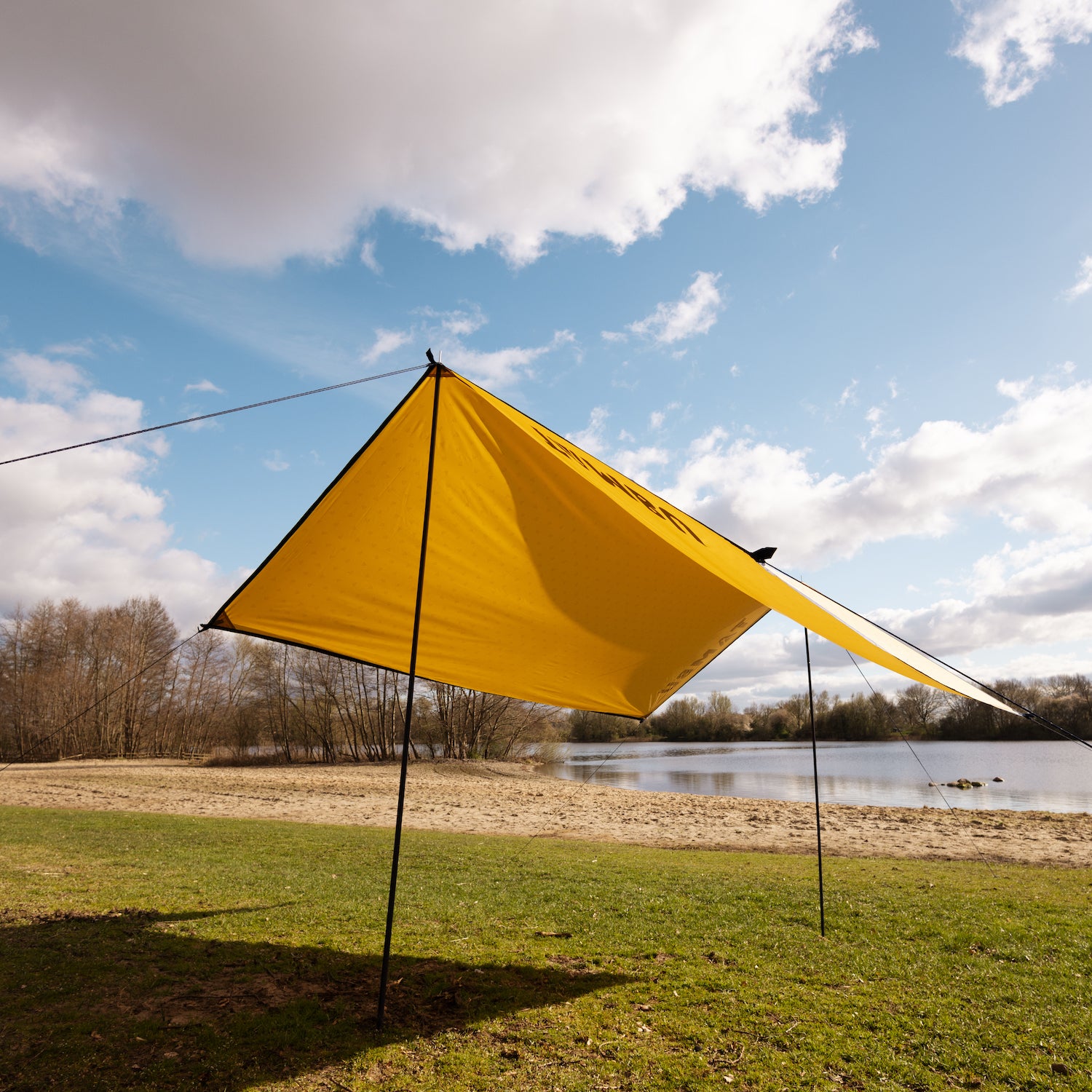 Gelbes multifunktionales Sonnensegel CHADA von Tambu Outdoor an einem Seeufer mit Sandstrand und Bäumen im Hintergrund