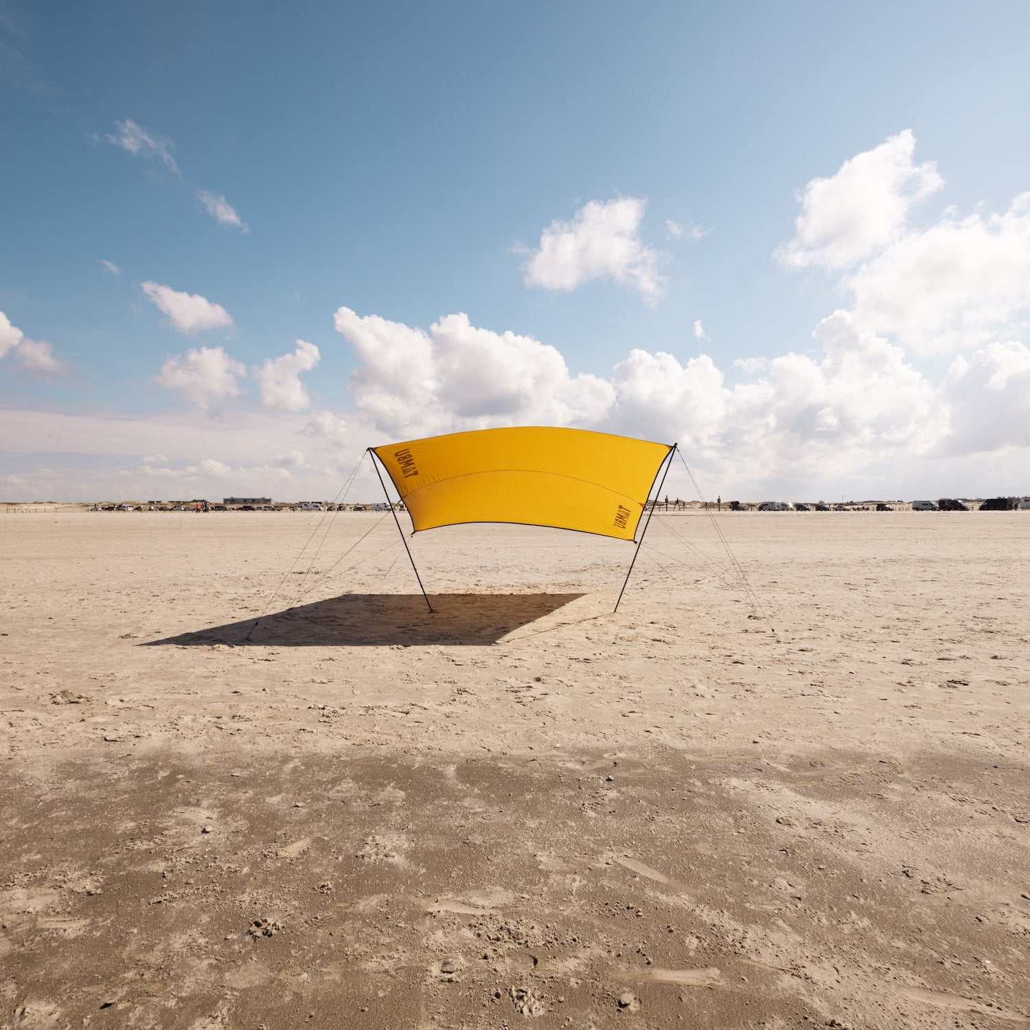Gelbes multifunktionales Sonnensegel CHADA von Tambu Outdoor auf sandigem Strand unter blauem Himmel mit Wolken