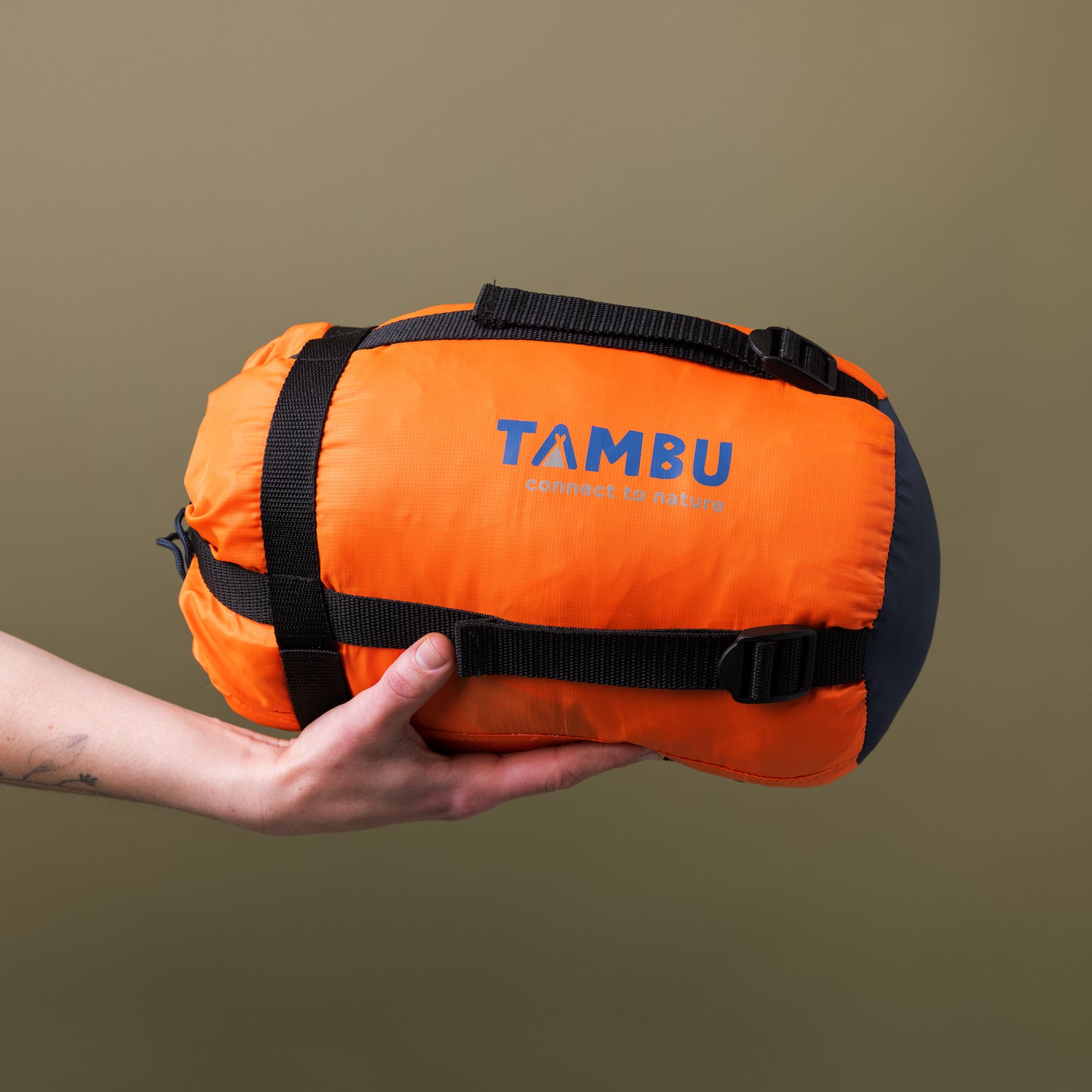 Hand hält kompakte, orange-schwarze Kompresstasche für Deckenschlafsack HASA 950 von Tambu Outdoor mit schwarzen Gurten und Logo 'TAMBU connect to nature'