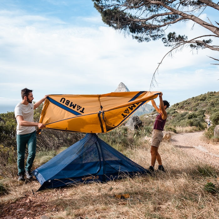 Zwei Personen bauen das orange-blaue KUTIR 2.0 Leichtgewichts-Trekkingzelt von Tambu Outdoor in einer bergigen Landschaft auf