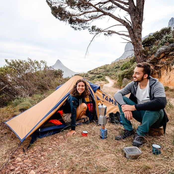 Zwei Personen vor dem KUTIR 2.0 Leichtgewichts-Trekkingzelt von Tambu Outdoor in der Natur, mit Campingkocher und Ausrüstung auf dem Boden