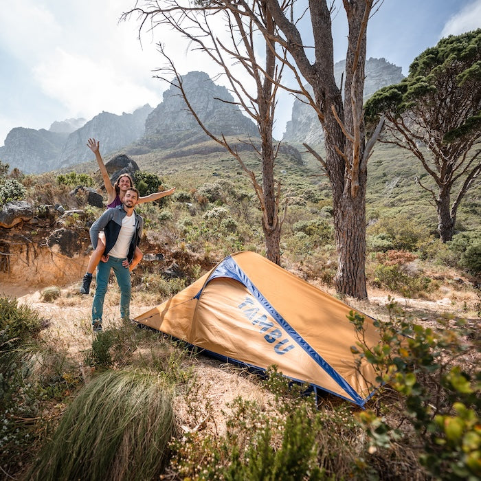 Leichtgewichts-Trekkingzelt KUTIR 2.0 von Tambu Outdoor in gelb mit blauem Logo, aufgebaut in bergiger Landschaft mit zwei fröhlichen Personen, eine trägt die andere auf dem Rücken