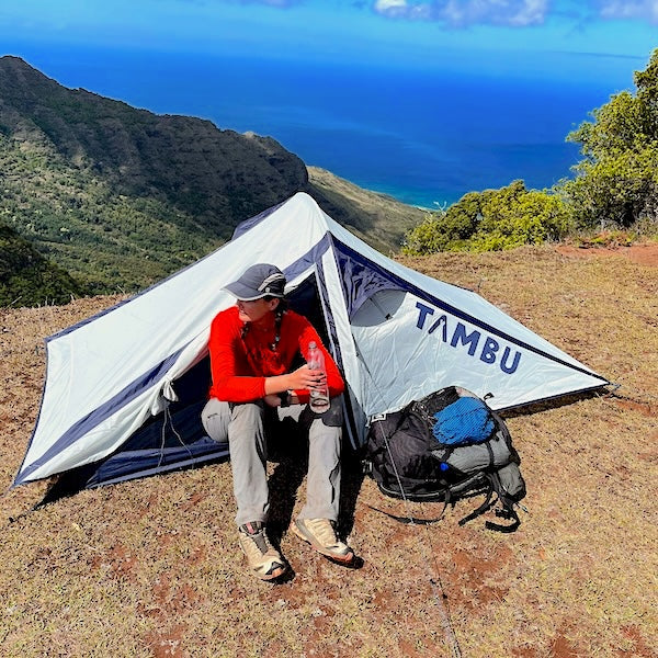 Person sitzt vor graublauem KUTIR Leichtgewichts-Trekkingzelt von Tambu Outdoor auf Berg mit Rucksack und Wanderstöcken