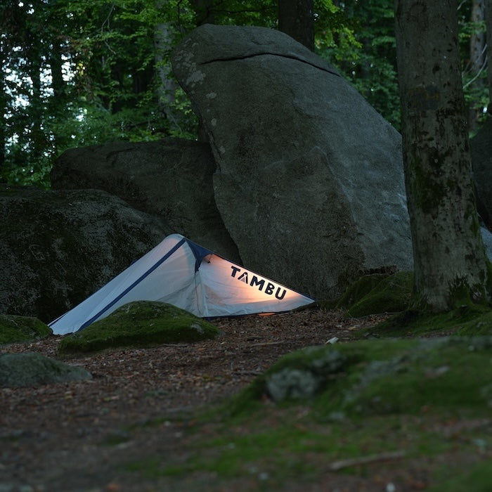 KUTIR 2-Personen Leichtgewichts-Trekkingzelt von Tambu Outdoor in Grau und Blau, beleuchtet im Wald neben großen Felsen