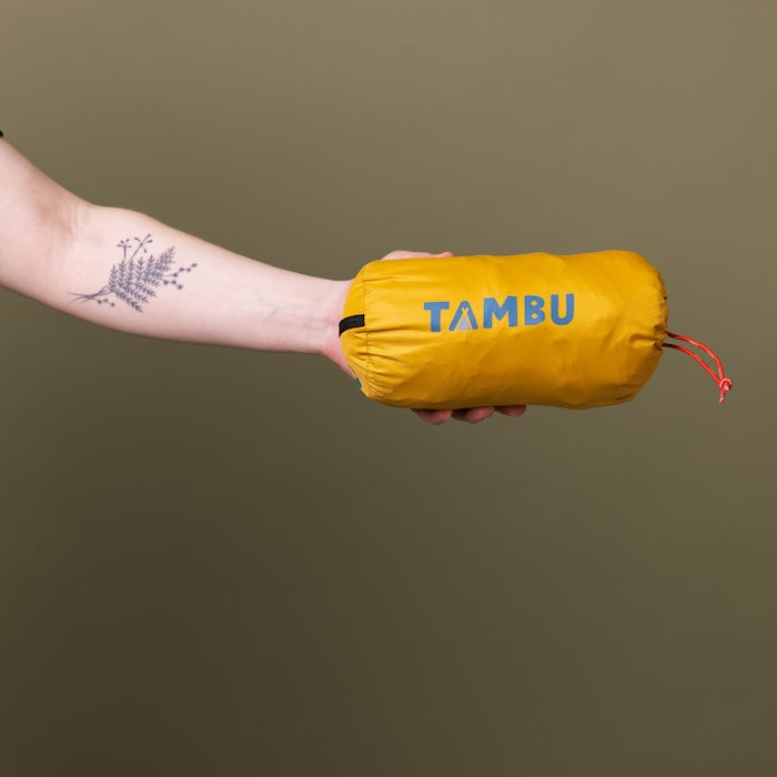 Hand hält gelben Biwacksack 350 von Tambu Outdoor mit grauem 'TAMBU' Schriftzug und rotem Kordelzug