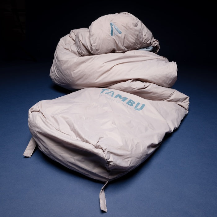 MOMI Mumienschlafsack 950 von Tambu Outdoor in hellgrauer Farbe mit Kapuze und blauem Innenfutter, liegend auf dunklem Untergrund