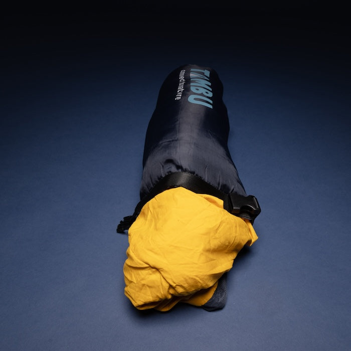 Mumienschlafsack NEEL 750 von Tambu Outdoor in dunkelblauem Packsack mit sichtbarem gelbem Innenfutter auf dunklem Hintergrund