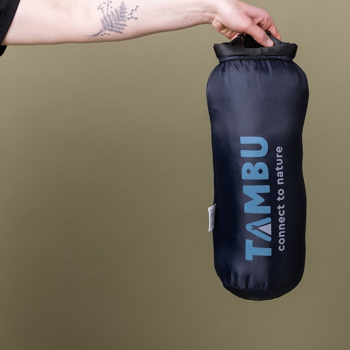 Hand hält kompakten dunkelblauen Packsack mit Aufschrift 'TAMBU connect to nature' für NEEL Mumienschlafsack 750 von Tambu Outdoor