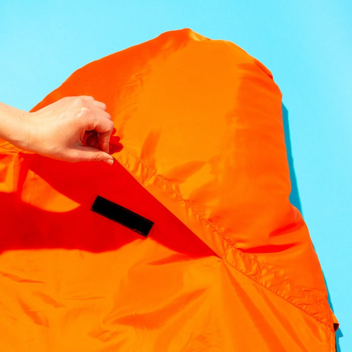Hand öffnet Klettverschluss an der Kapuze des orangefarbenen OFRA 800 Mumienschlafsacks von Tambu Outdoor vor blauem Hintergrund