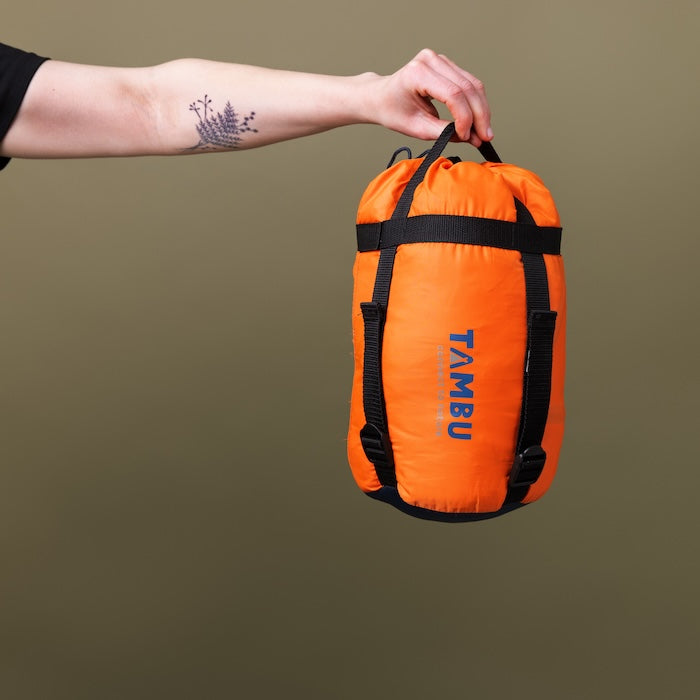 Hand hält orangefarbenen Packsack mit schwarzem Tragegriff und Aufschrift 'TAMBU' für OFRA 800 Mumienschlafsack von Tambu Outdoor