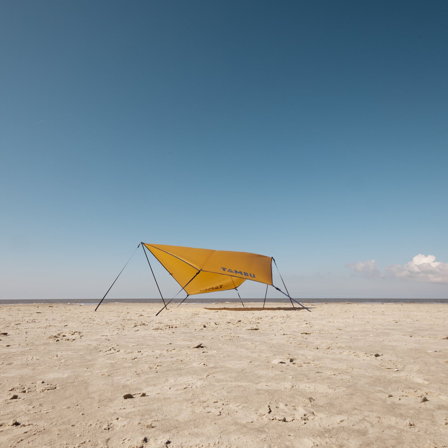 Gelbes PALA Sonnensegel von Tambu Outdoor mit schwarzen Aufstellstangen auf sandigem Strand unter blauem Himmel mit Wolken