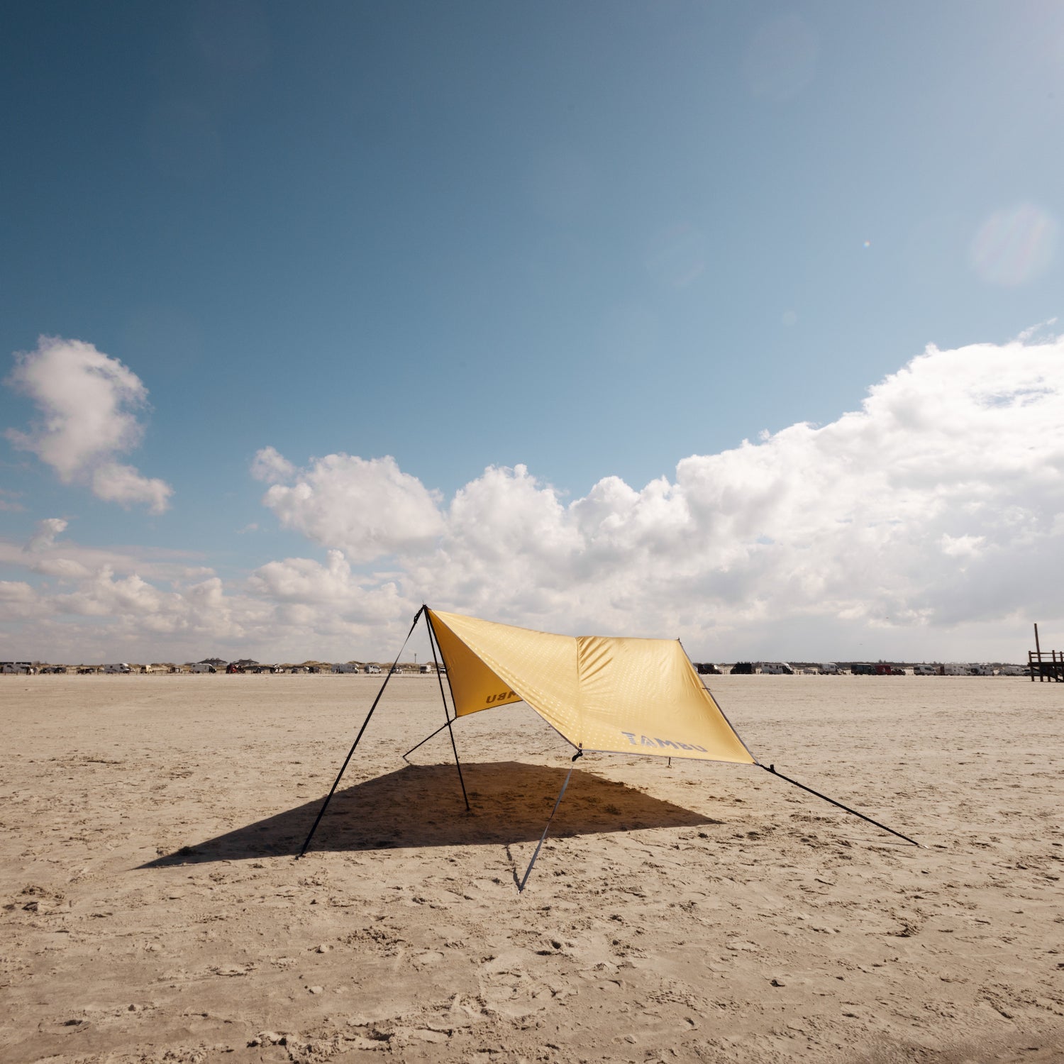 Gelbes PALA Sonnensegel von Tambu Outdoor mit schwarzen Aufstellstangen auf sandigem Strand unter blauem Himmel mit Wolken