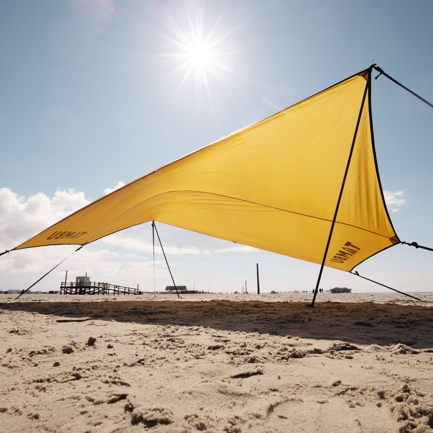 Gelbes Sonnensegel PALA von Tambu Outdoor mit schwarzen Aufstellstangen am sonnigen Strand gespannt