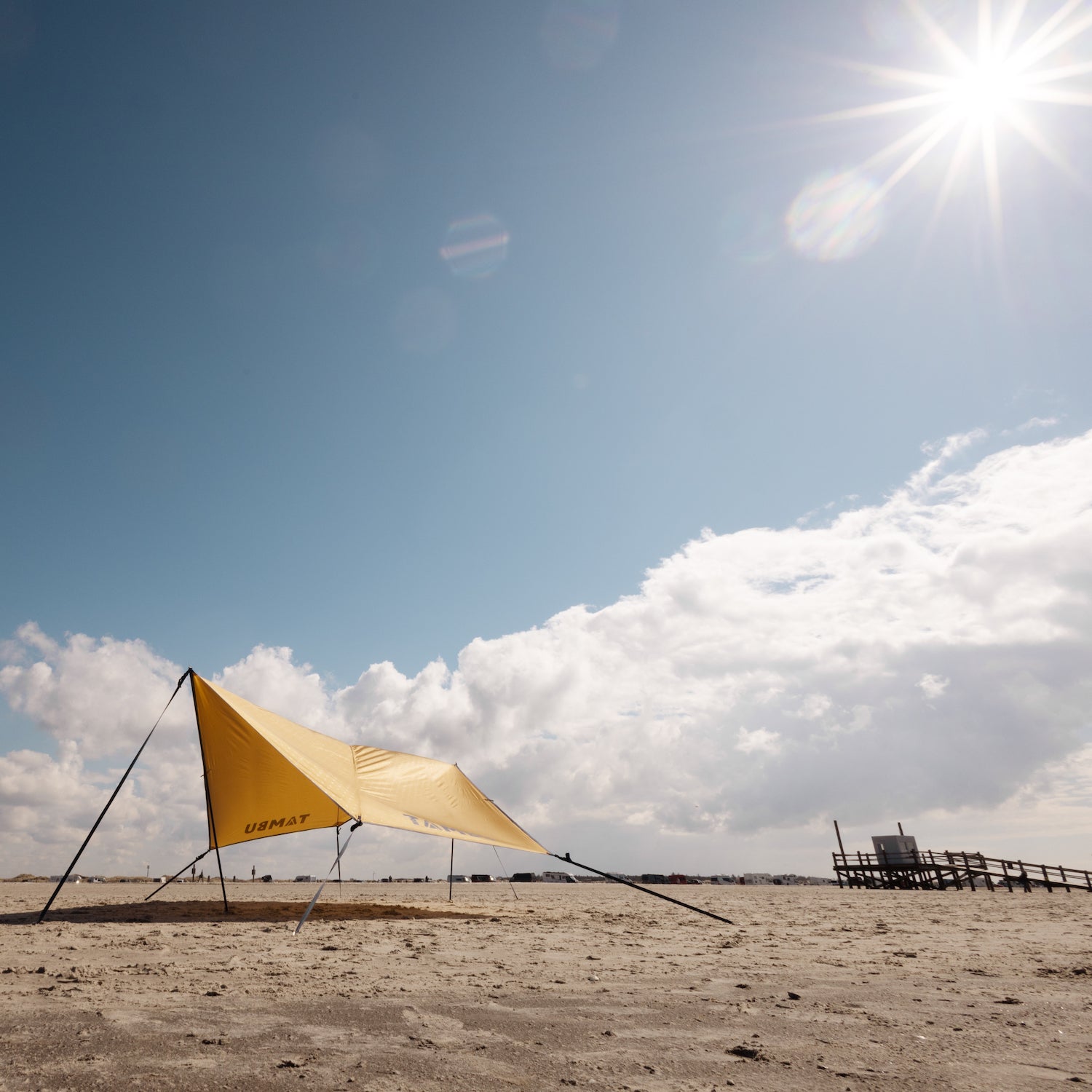 Gelbes PALA Sonnensegel mit schwarzen Aufstellstangen von Tambu Outdoor auf sandigem Strand unter blauem Himmel mit Sonne und Wolken