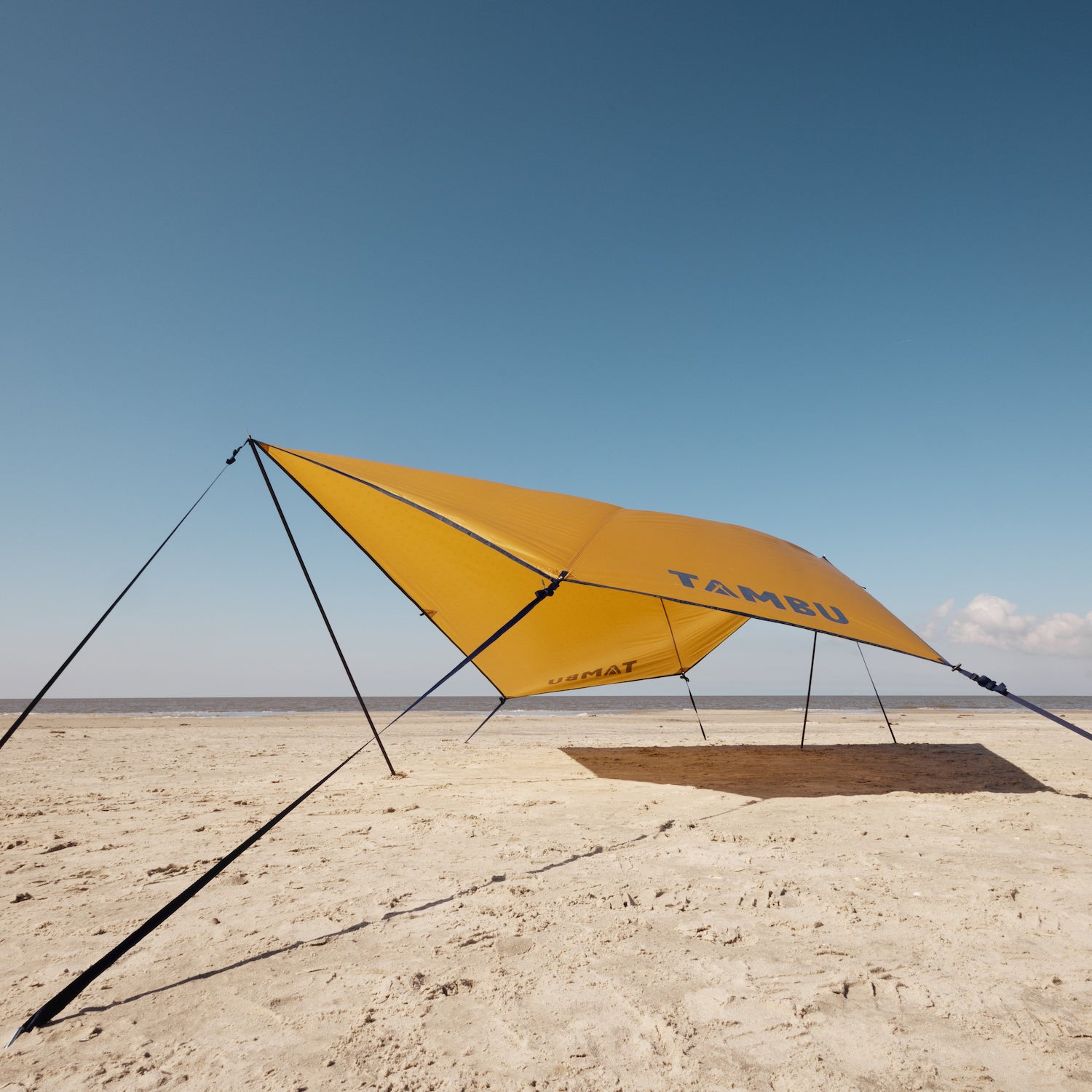 Gelbes PALA Sonnensegel von Tambu Outdoor mit schwarzen Aufstellstangen auf sandigem Strand unter blauem Himmel mit Wolken