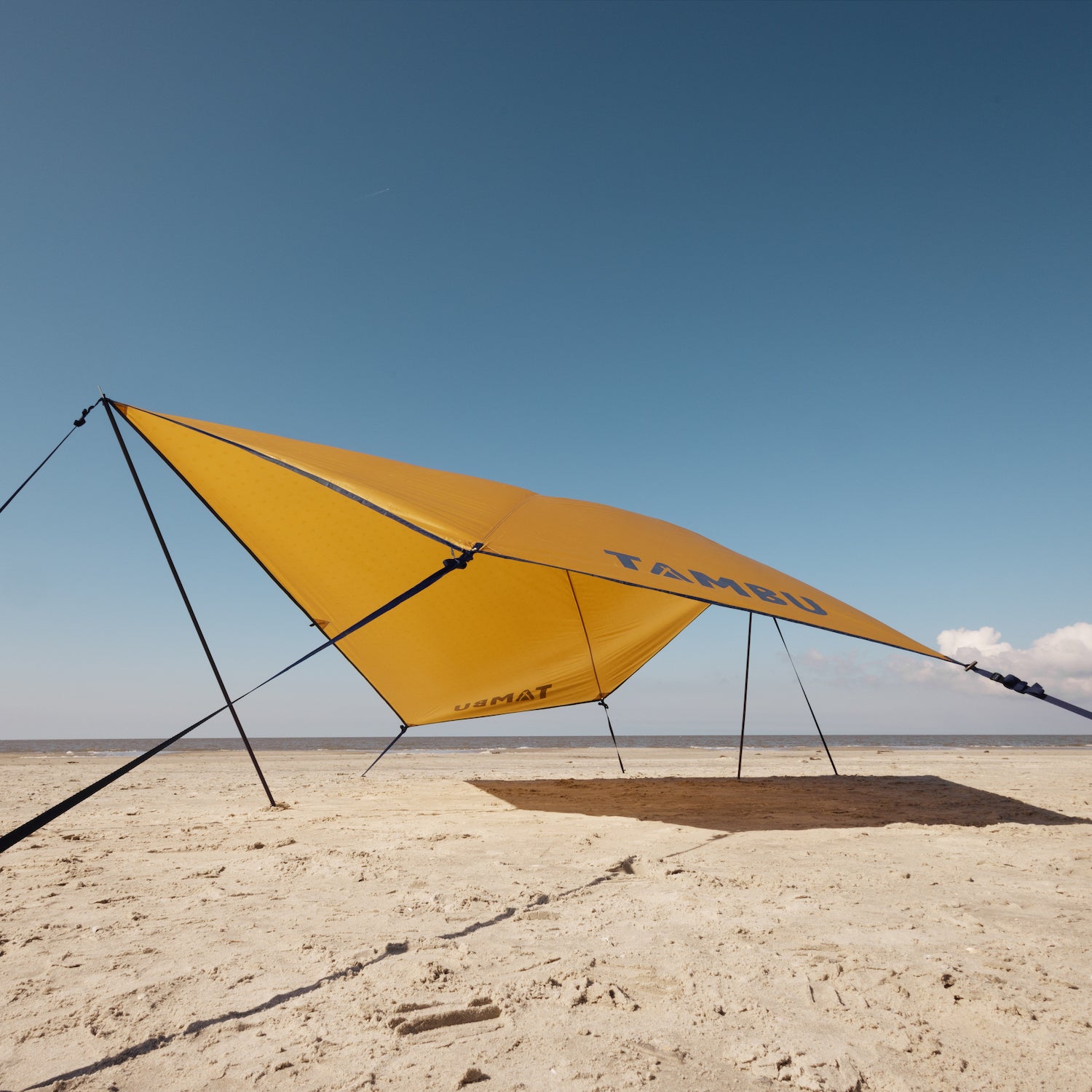 Gelbes PALA Sonnensegel von Tambu Outdoor mit schwarzen Aufstellstangen auf sandigem Strand unter blauem Himmel mit Wolken