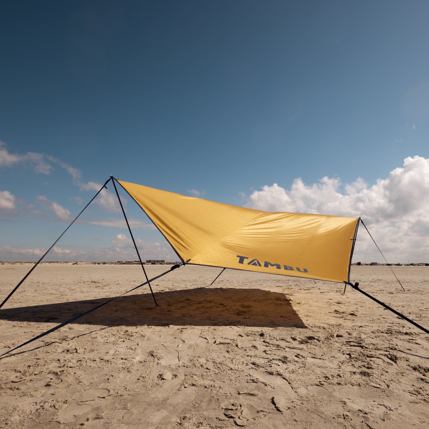 Gelbes Sonnensegel mit Aufstellstangen von Tambu Outdoor am Strand, mit dem Schriftzug 'TAMBU' auf dem Segel