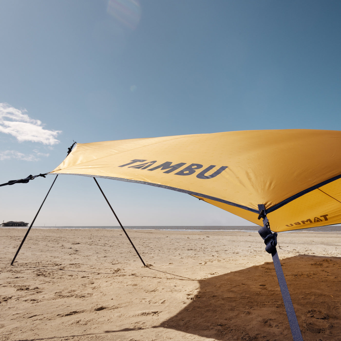 Gelbes Sonnensegel mit Aufstellstangen von Tambu Outdoor am Strand, mit dem Schriftzug 'TAMBU' auf dem Segel