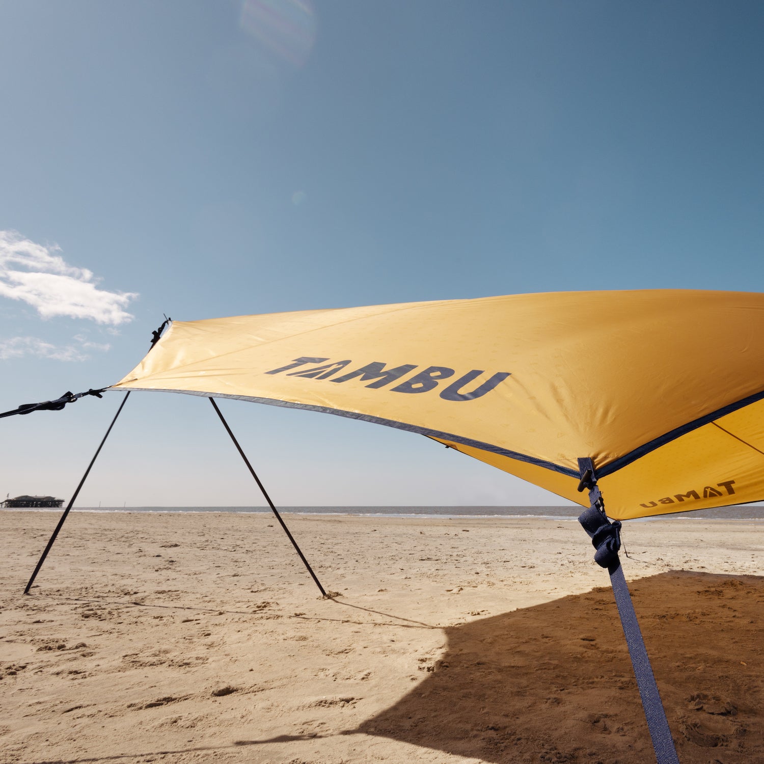 Gelbes Sonnensegel mit Aufstellstangen von Tambu Outdoor am Strand, mit dem Schriftzug 'TAMBU' auf dem Segel
