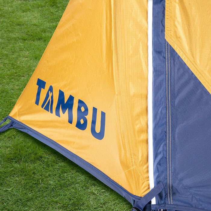 Ecke des gelb-blauen SURANGA 2.0 Leichtgewichts-Trekkingzelts von Tambu Outdoor auf grünem Gras mit blauem Tambu-Logo