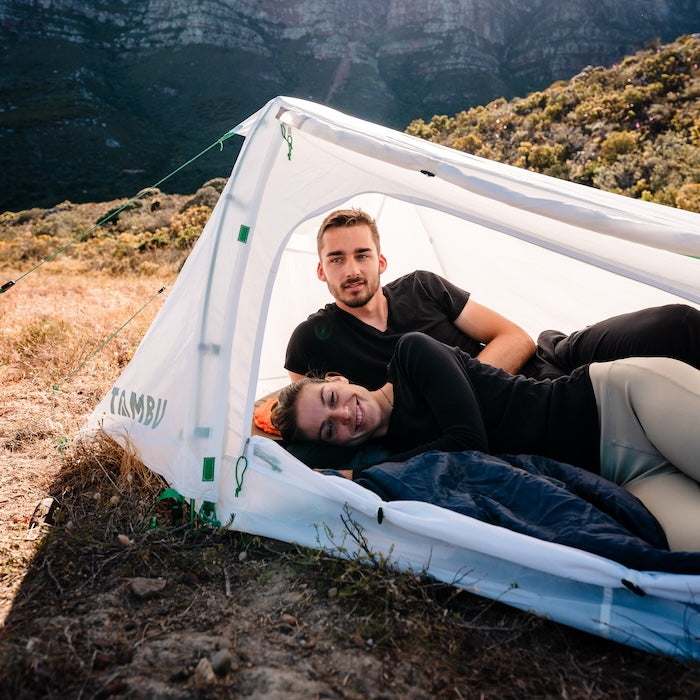 Zwei Personen liegen entspannt im weißen SURAN 2-Personen Trekking-Tunnelzelt von Tambu Outdoor in einer bergigen Landschaft