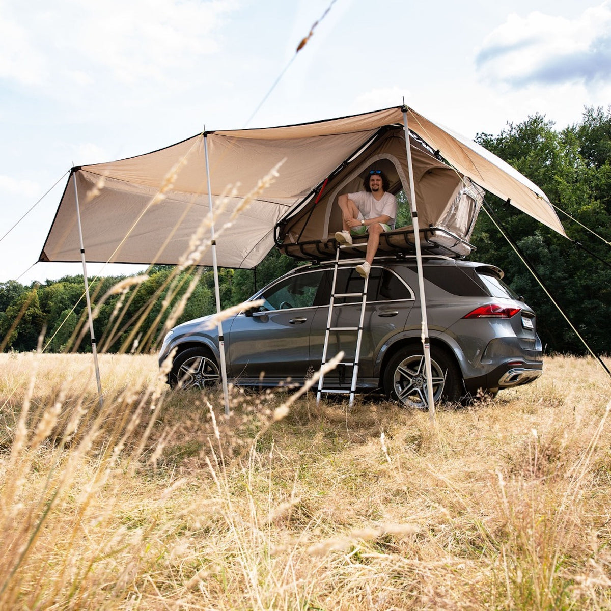 Graues SUV mit Dachzelt und YANO Sonnensegel von Tambu Outdoor, Mann sitzt entspannt im geöffneten Zelt, umgeben von trockenem Gras und Bäumen