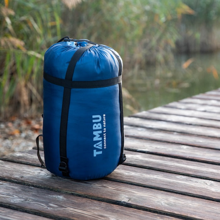 Blauer Packsack mit Aufschrift 'TAMBU connect to nature' steht auf einem Holzsteg am See vom Mumienschafsack TALU 1500