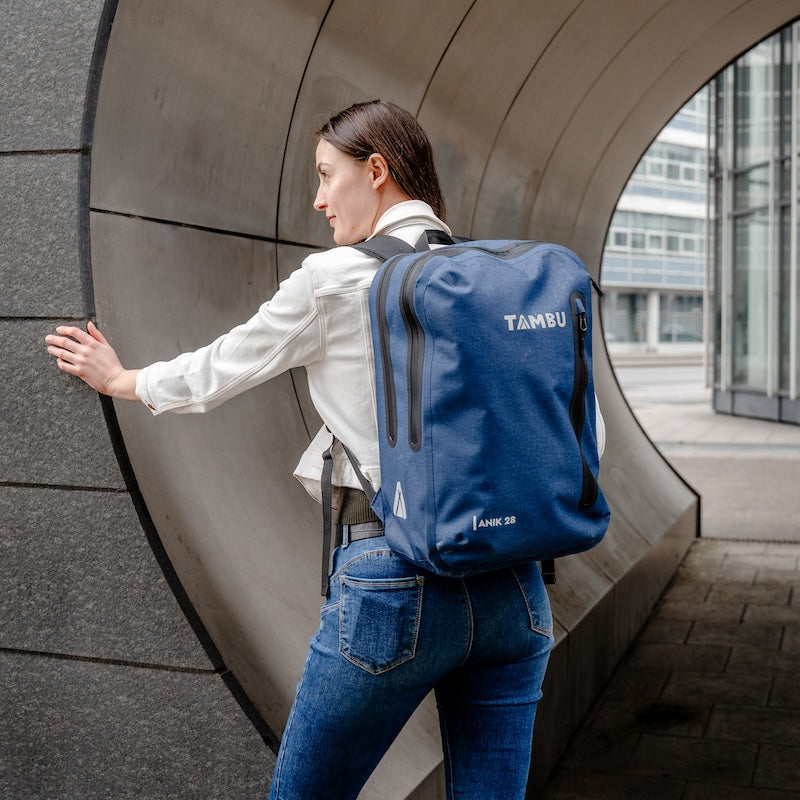 Frau mit weißem Hemd und Jeans trägt einen blauen wasserabweisenden ANIK 28 Rucksack von Tambu Outdoor in urbaner Umgebung