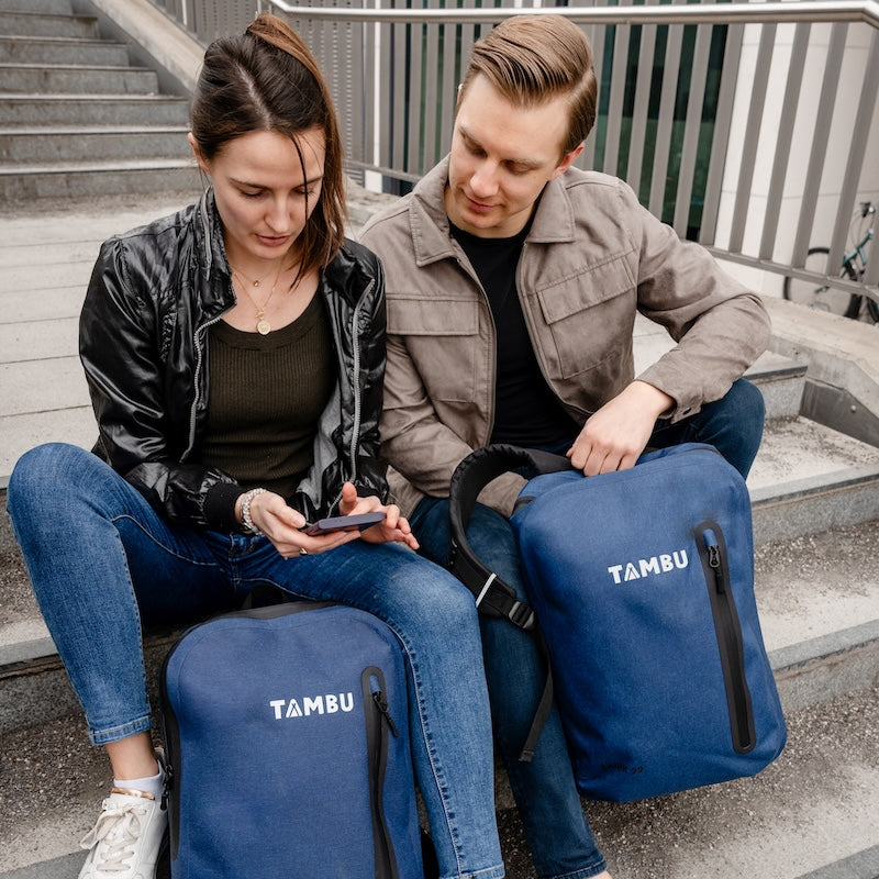 Junge Frau und Mann sitzen auf Treppen, beide mit dunkelblauen wasserabweisenden ANIK 22 oder 28 Rucksäcken von Tambu Outdoor, schauen gemeinsam auf ein Smartphone
