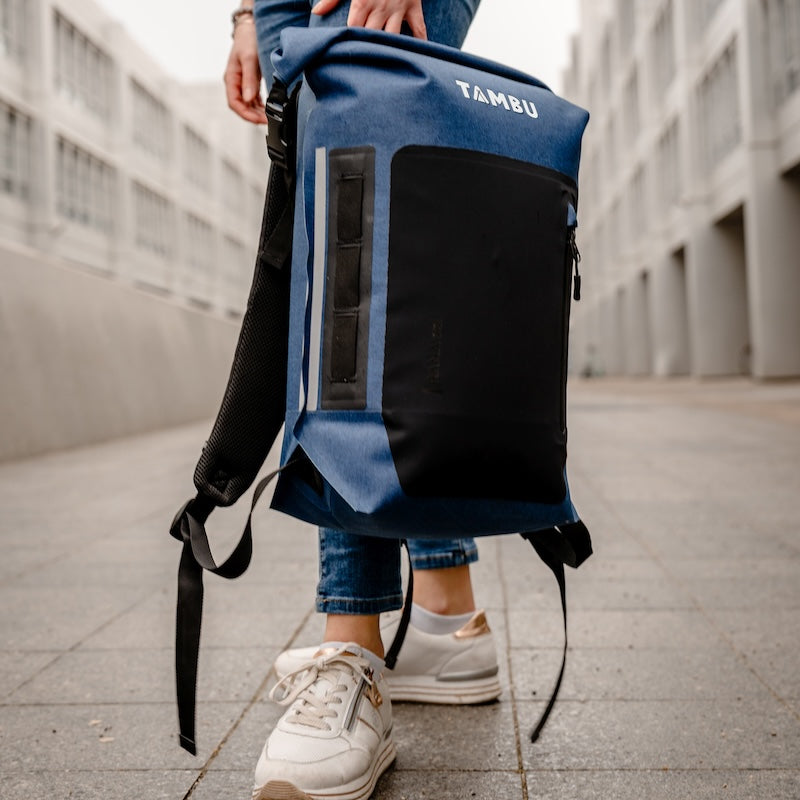 Person hält einen blauen RANA 22 Roll-on-Top Rucksack von Tambu Outdoor, wasserabweisend, mit schwarzer Fronttasche und reflektierendem Streifen