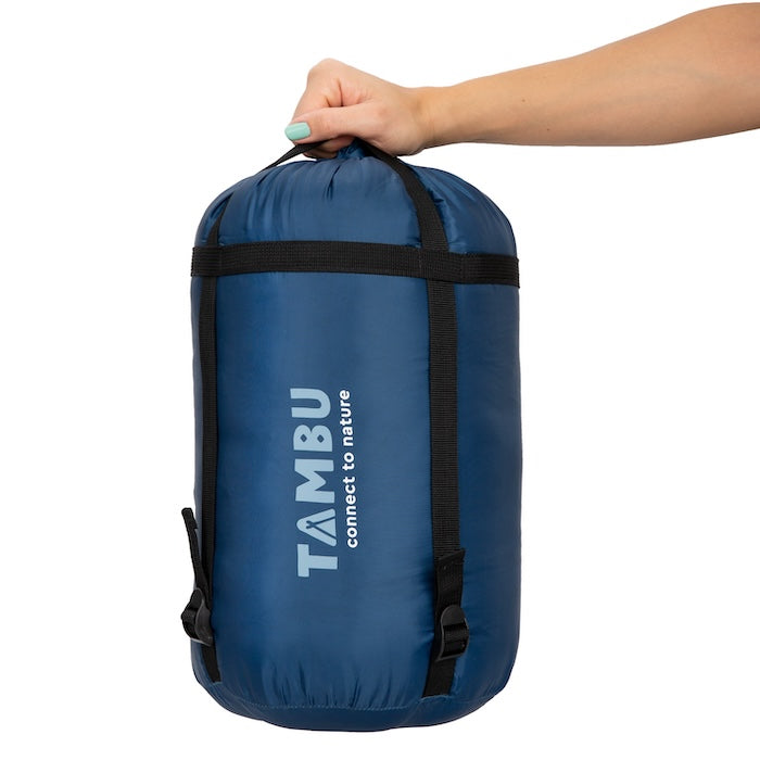 Hand hält kompakten blauen Mumienschlafsack TALU 1500 von Tambu Outdoor im Packsack mit schwarzem Tragegurt und Aufschrift 'TAMBU connect to nature'