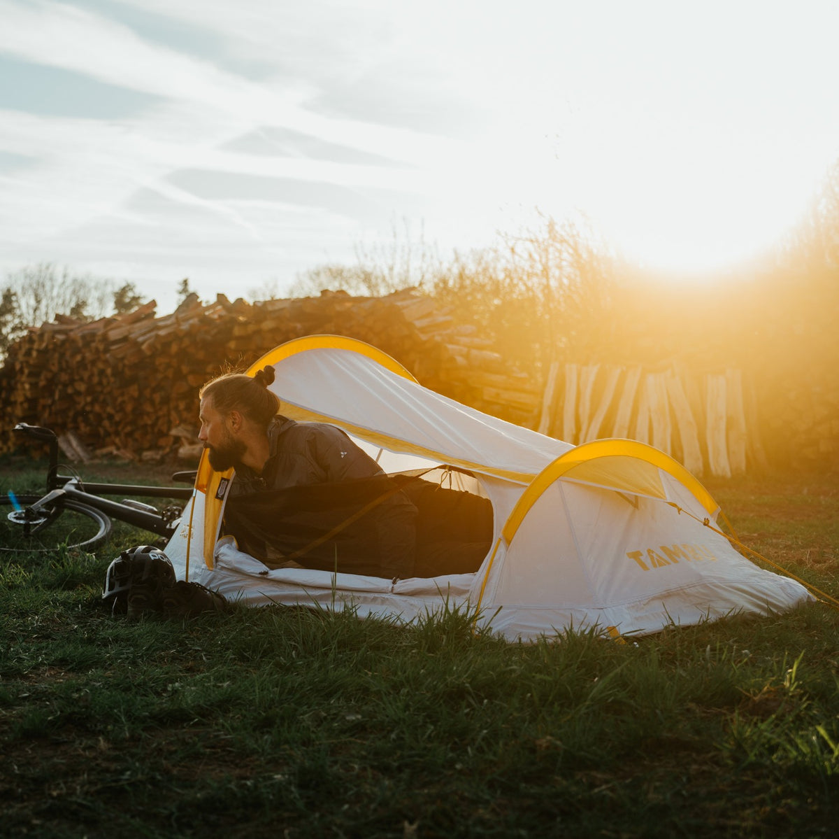 Person liegt im weißen und gelben ROBI Leichtgewichtszelt von Tambu Outdoor auf einer Wiese bei Sonnenuntergang