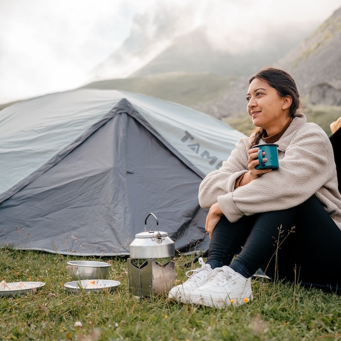 Frau sitzt auf Gras vor grauem TIKONA  Trekking Kuppelzelt von Tambu Outdoor, hält blaue Tasse, Campinggeschirr und Kaffeekanne stehen daneben