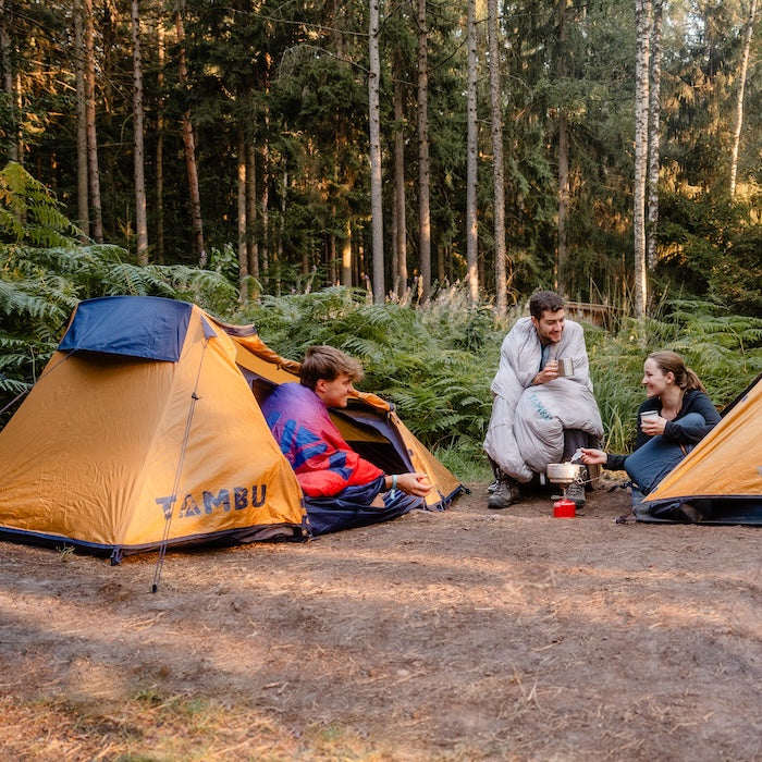 Zwei gelbblaue SURANGA 2.0 Leichtgewichts-Trekkingzelte von Tambu Outdoor im Wald, drei Personen sitzen draußen, kochen und trinken aus Bechern